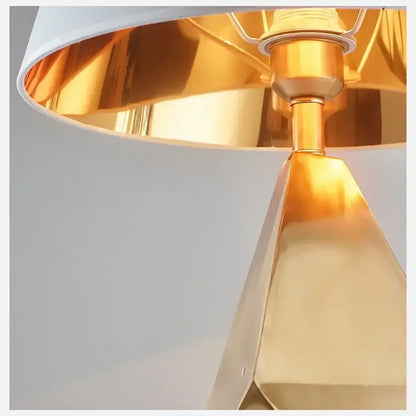 Modern gold geometric table lamp with a sleek three-dimensional base and white shade with interior gold lining for bedroom use