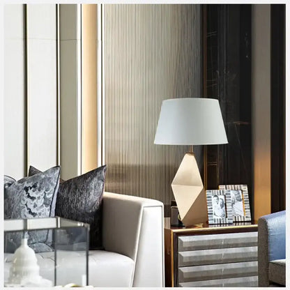 Modern gold geometric table lamp with a white fabric shade and a faceted three-dimensional geometric base for bedroom use