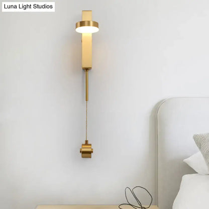 Modern gold metal wall sconce with cylindrical light head and pull-chain switch emitting warm white light for bedroom use