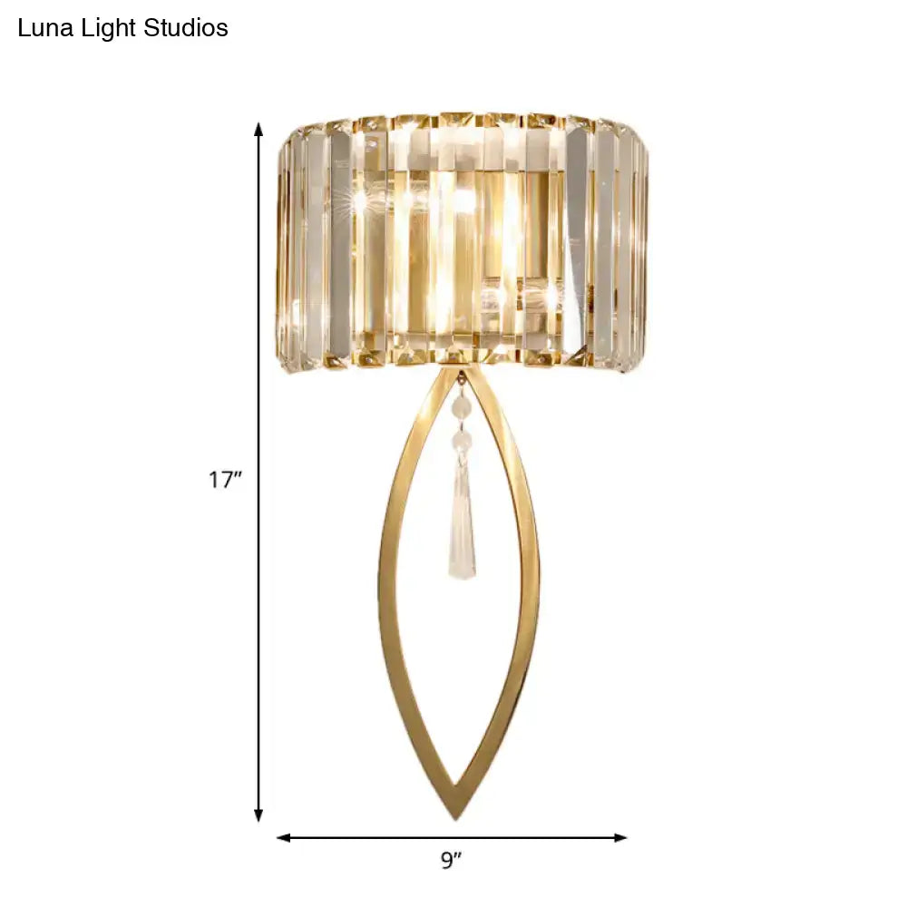 Modern gold metallic drum shade wall light with a cylindrical crystal shade and elongated leaf-shaped frame for living room
