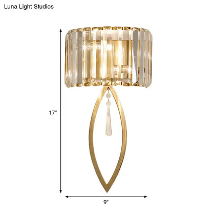Modern gold metallic drum shade wall light with a cylindrical crystal shade and elongated leaf-shaped frame for living room