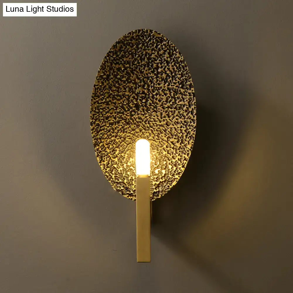 Modern gold oval metal wall sconce with textured hammered finish and sleek vertical stem for bedroom lighting