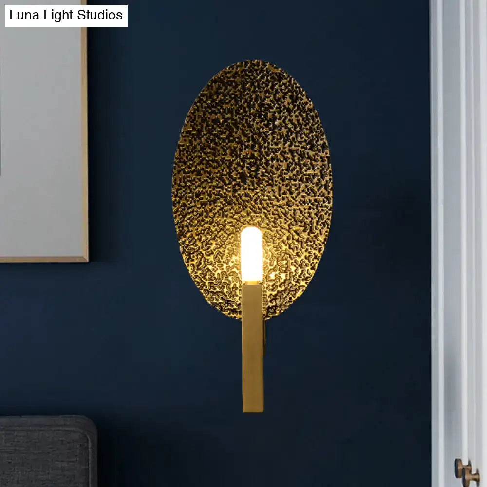 Modern gold oval metal wall sconce with textured hammered face and sleek vertical stem for bedroom lighting