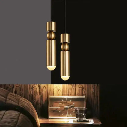 Two modern decorbites gold tube pendant lights with cylindrical and spherical brass designs hang above a wooden nightstand