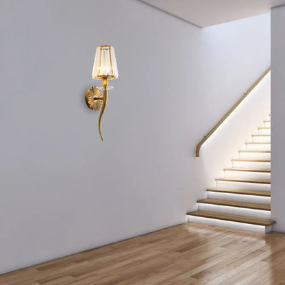 Modern gold wall light with a clear tapered shade and gold-toned curved arm for stairways and hallways