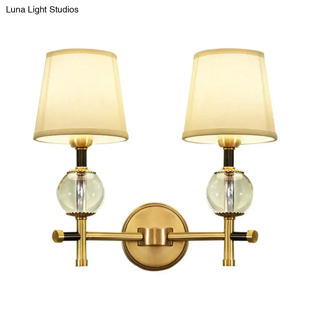 Brass-finished modern gold wall light with two clear crystal globes and cream conical fabric shades for bedroom use