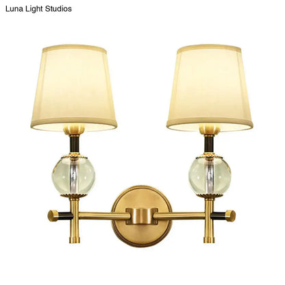 Brass-finished modern gold wall light with two clear crystal globes and cream conical fabric shades for bedroom use