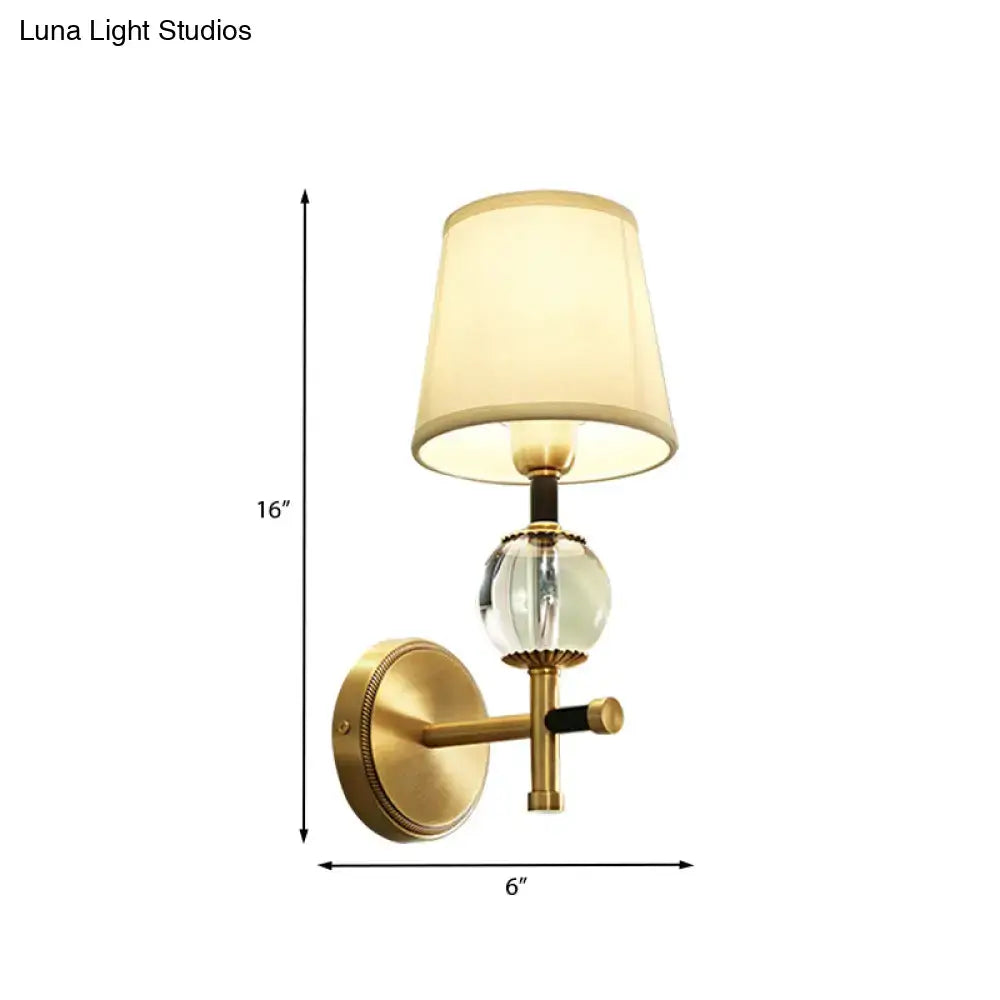 Modern gold wall light featuring a gold-toned brass arm, clear crystal globe, and off-white conical fabric shade for bedroom use