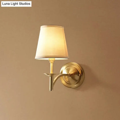Brass-finished modern wall light with tapered off-white fabric shade and compact fixture depth for bedroom sconce lighting