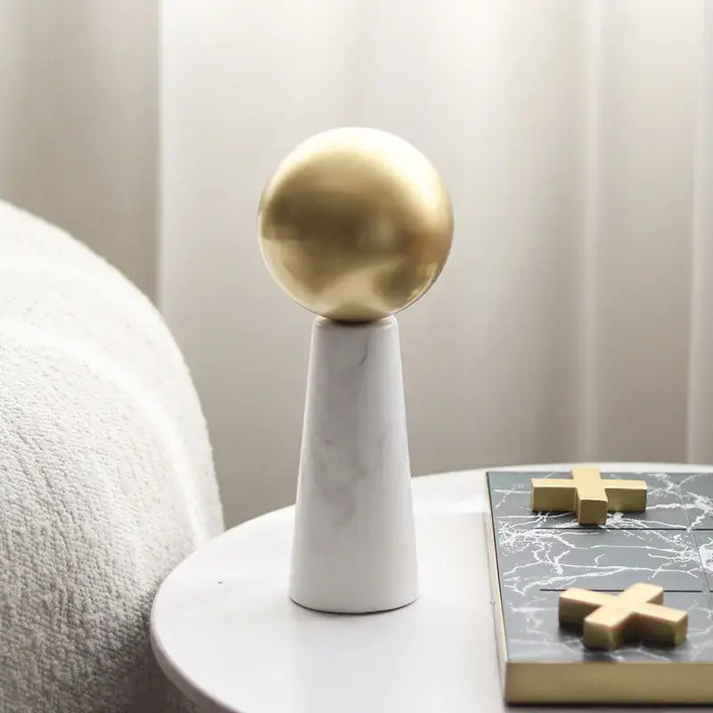 A modern golden ball spherical ornament with a polished finish on a white marble cone base for living room decor