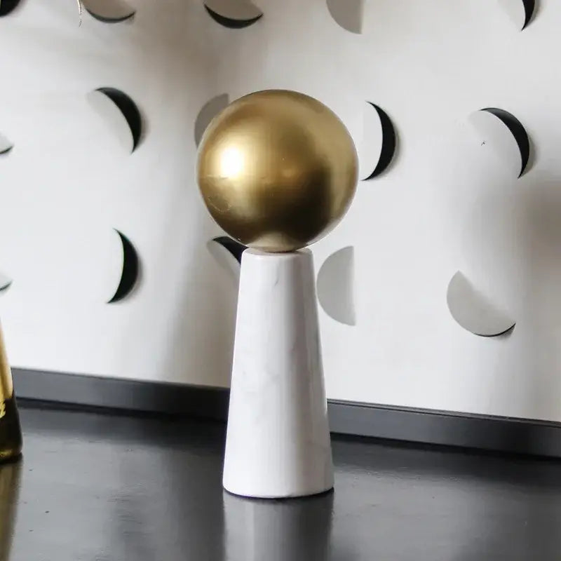 A sleek golden ball rests on a white marble cone pedestal as a modern living room spherical ornament