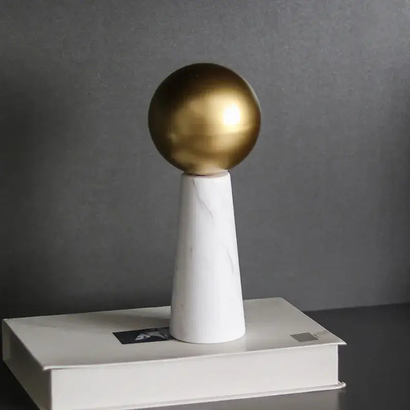 A sleek polished golden ball resting on a white marble cone pedestal modern spherical ornament for living room