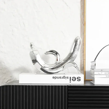 Shiny chrome abstract sculpture with flowing curved forms resting on a white book in modern golden banana figurine decor
