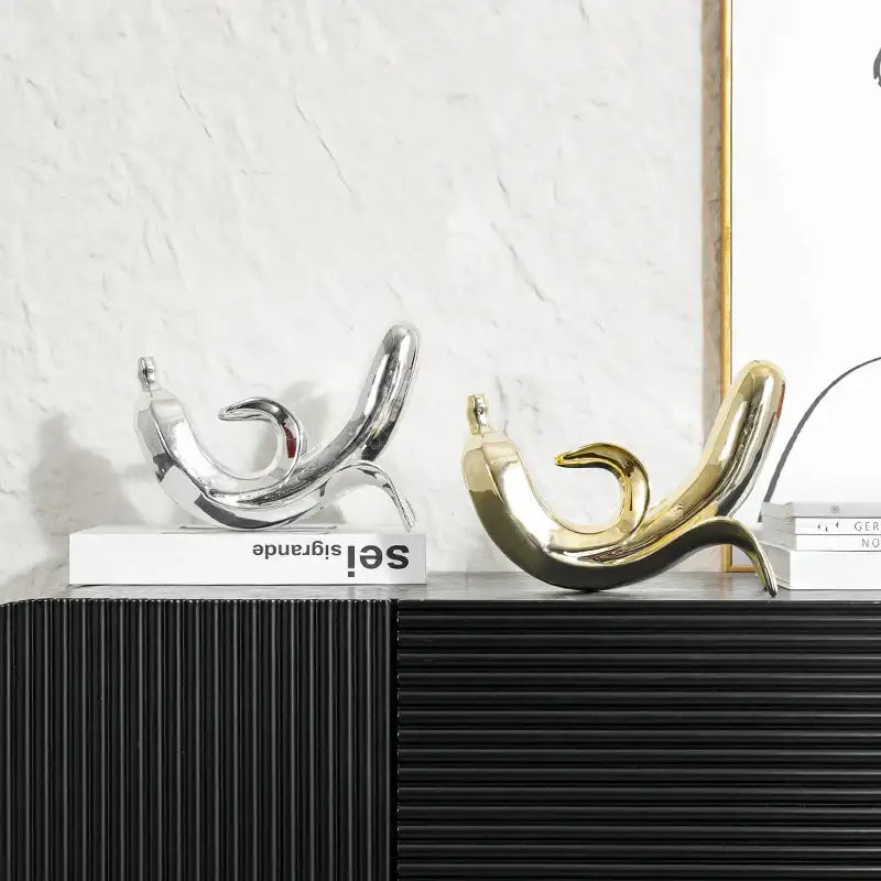 Two sleek abstract golden banana figurines in silver and gold rest elegantly on books on a black ribbed surface