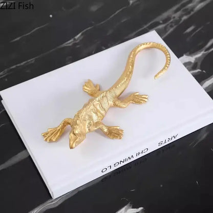 Gold-colored metal golden lizard sculpture with textured scales and curved tail resting on a white book for living room decor