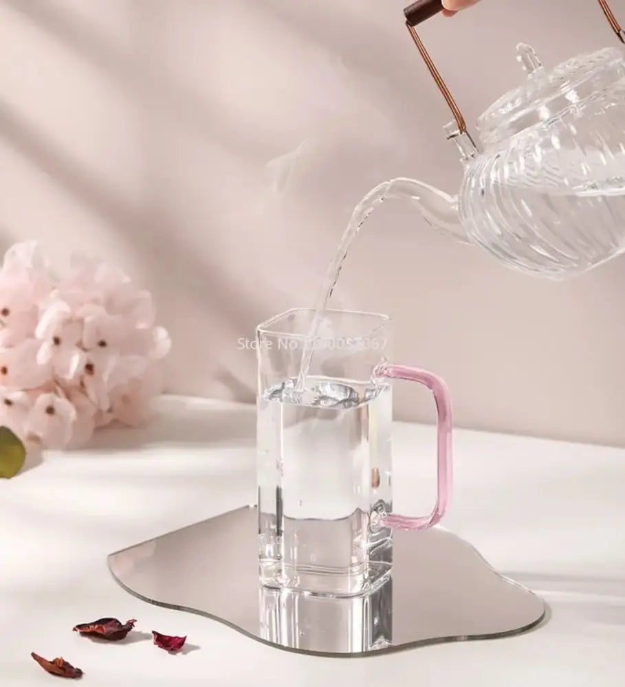 Clear glass teapot pours steaming water into a delicate pink handled glass mug on an elegant mirrored tray with square glass mug