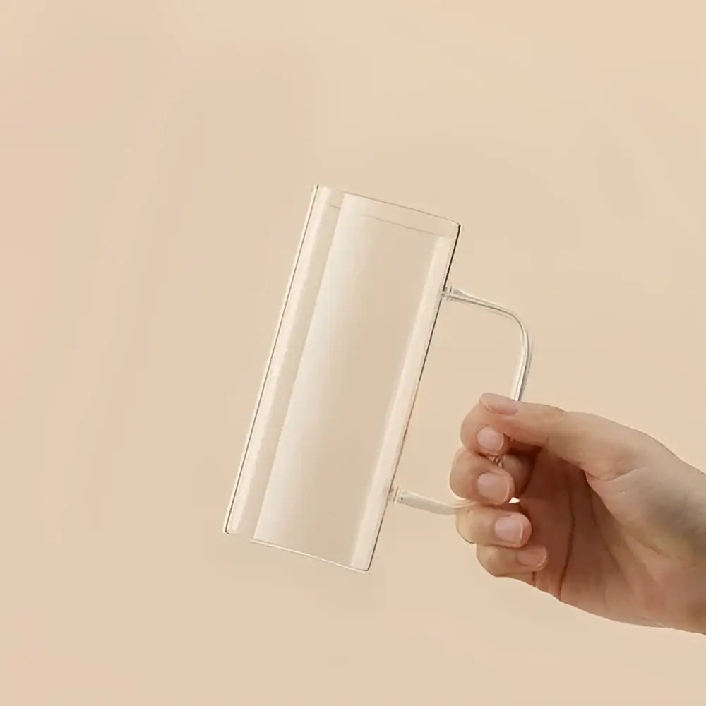 Clear glass square glass mug with modern green minimalist handle held in hand against soft beige background