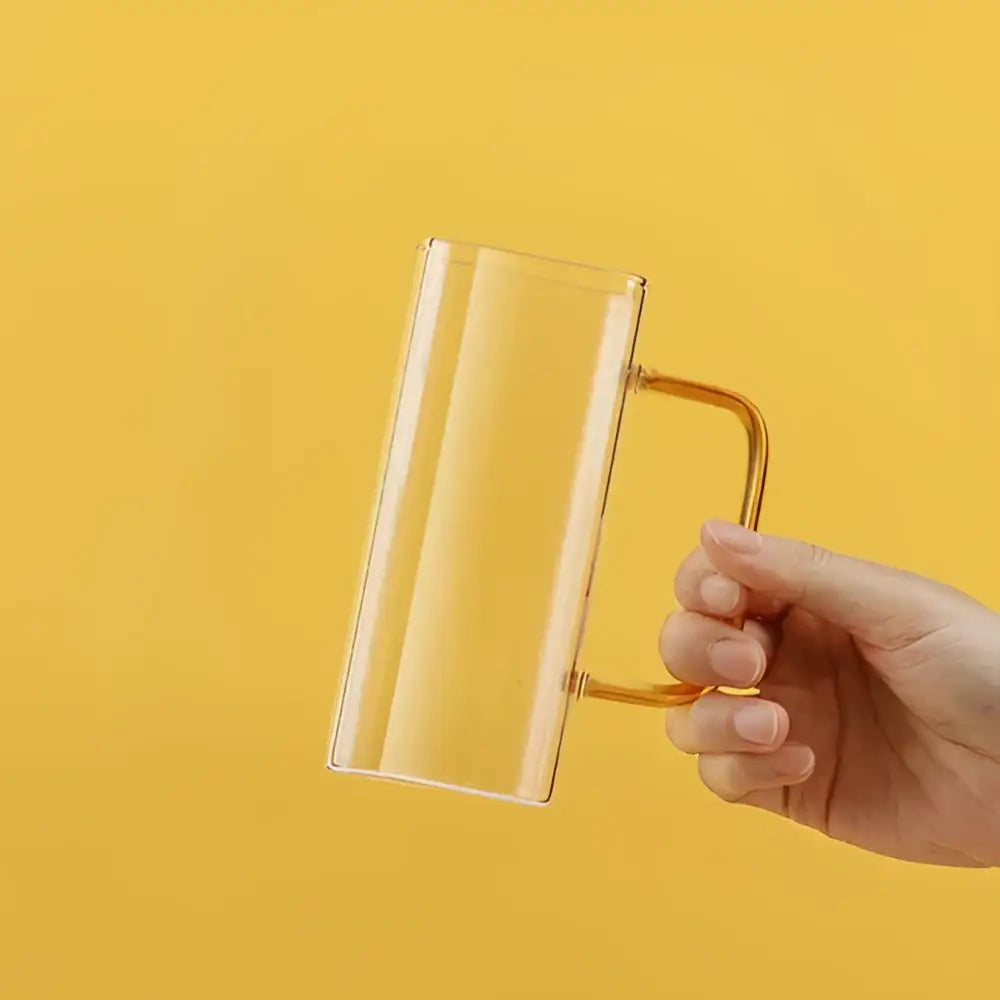 Clear glass mug with gold-toned handle held in hand against yellow background showcasing a modern square glass mug