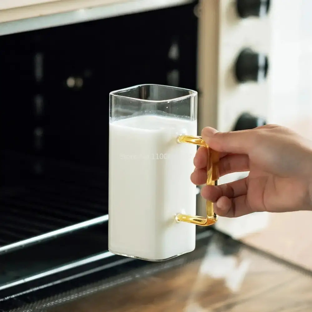 A clear square glass mug with a gold metal handle filled with milk for modern kitchen drinking use