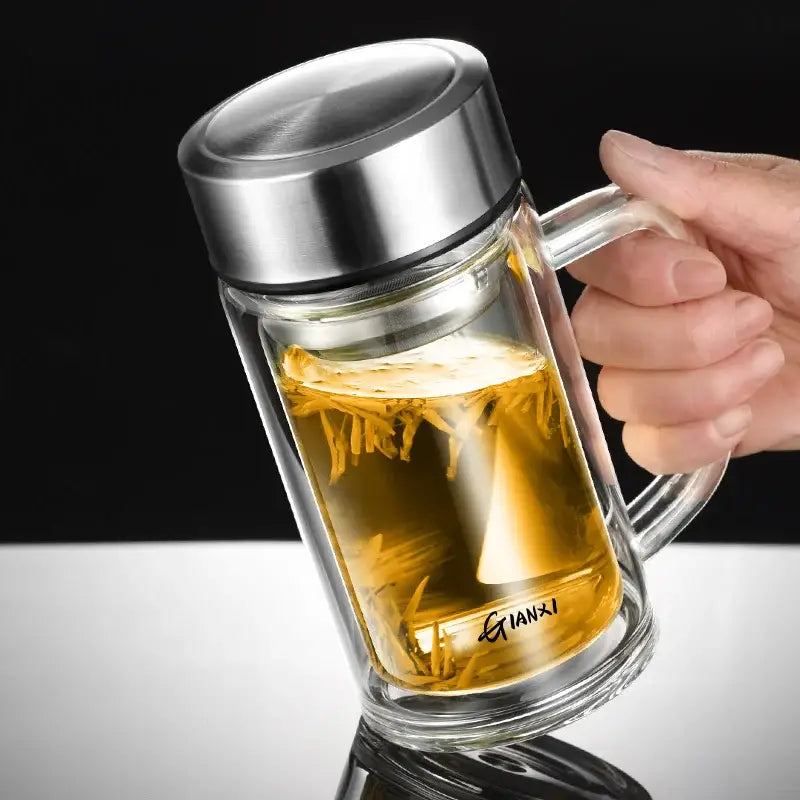 Modern insulated clear double-walled glass tea cup with stainless steel lid and handle filled with golden tea leaves