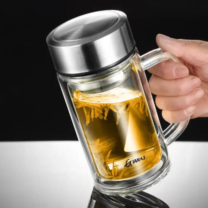 Modern insulated clear double-walled glass tea cup with stainless steel lid and handle filled with golden tea leaves