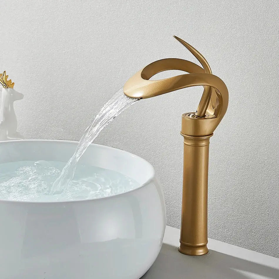Golden waterfall basin faucet with sleek curved design flowing water into white ceramic sink for modern bathroom vanity
