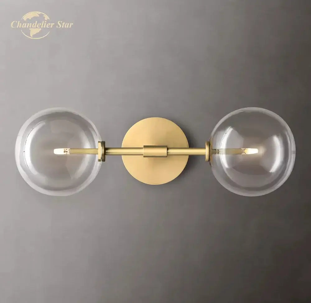 Modern metallic gold-toned wall sconce with two clear glass globe shades featuring a minimalist design for bedroom lighting
