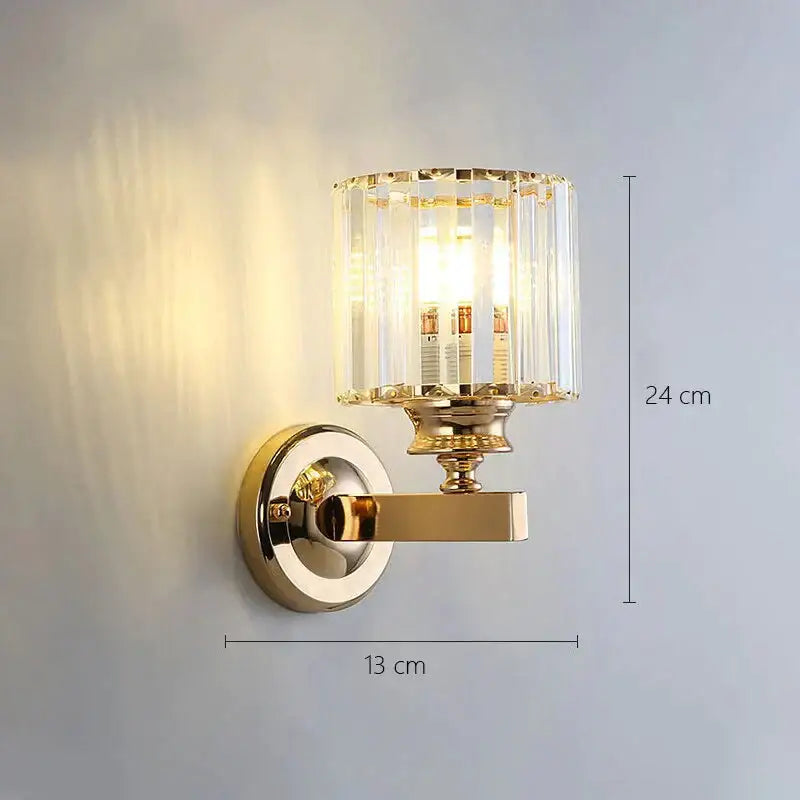 Modern minimalist wall lamp gold toned with cylindrical crystal glass shade and ornate metal bracket for bedroom use