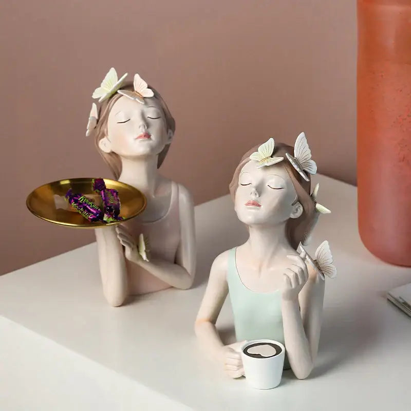 Two modern pink butterfly girl resin figures with butterfly hair accessories holding a gold tray and white mug for decor