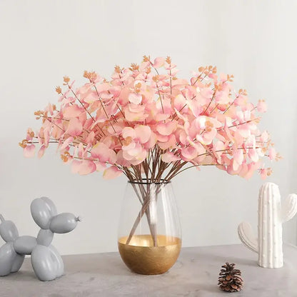 A clear glass vase with a gold-finished base holds a modern pink eucalyptus flower arrangement for living room decor