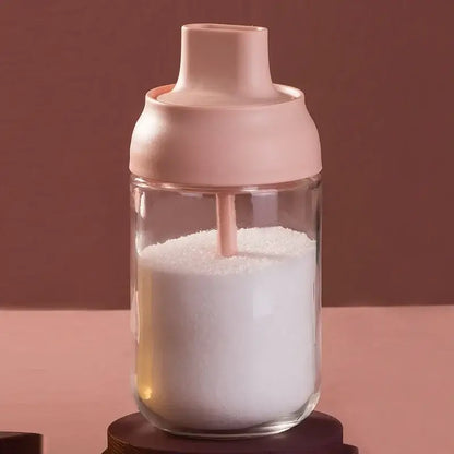 A clear glass seasoning bottle with a soft pink lid and straw filled with white granulated sugar for kitchen storage