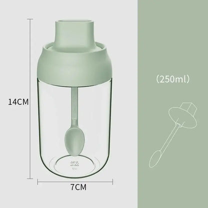 A clear glass seasoning bottle with a matte sage green lid and integrated spoon for convenient kitchen use