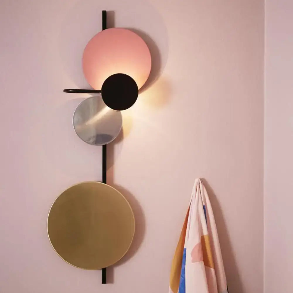 Modern geometric wall sconce featuring stacked circular discs in matte pink, black, mirrored silver, and brushed gold tones mounted on a sleek black vertical rod.