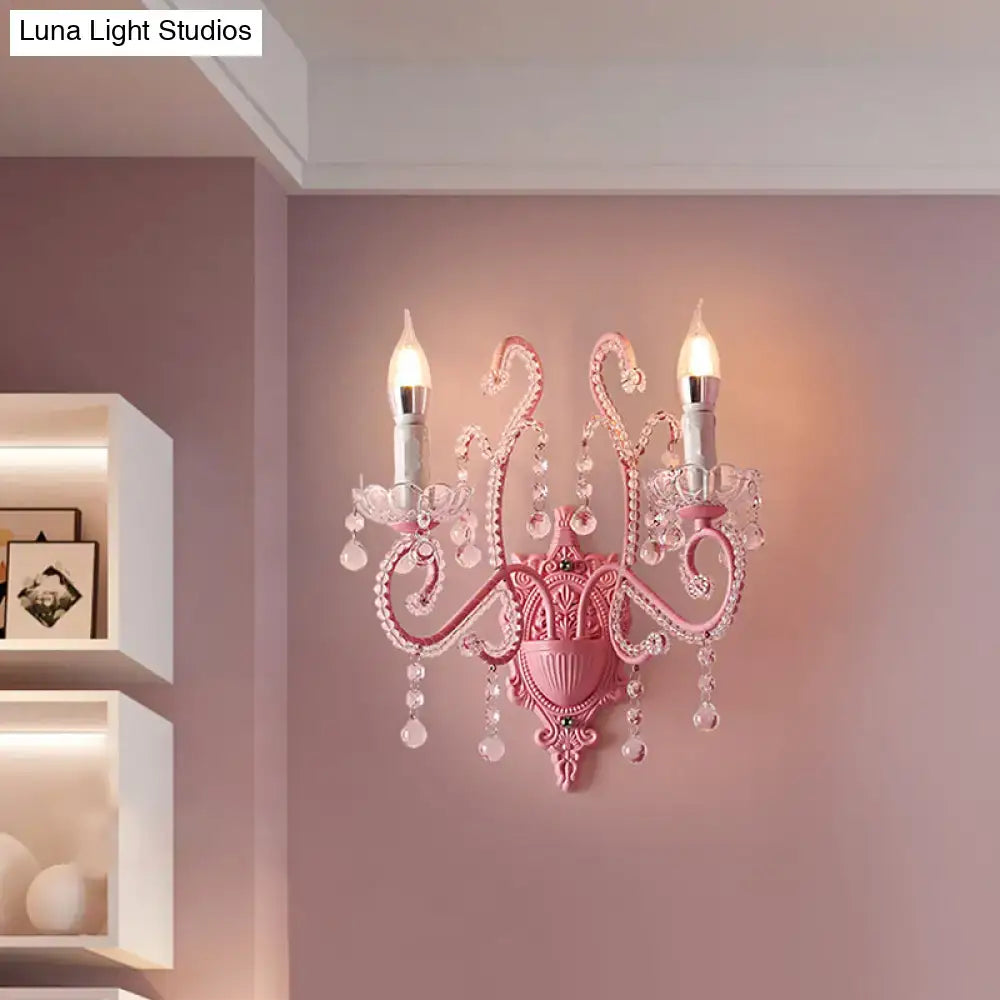 Modern pink metallic scroll arm wall lamp with crystal droplets and two lit candle-style bulbs for bedroom use