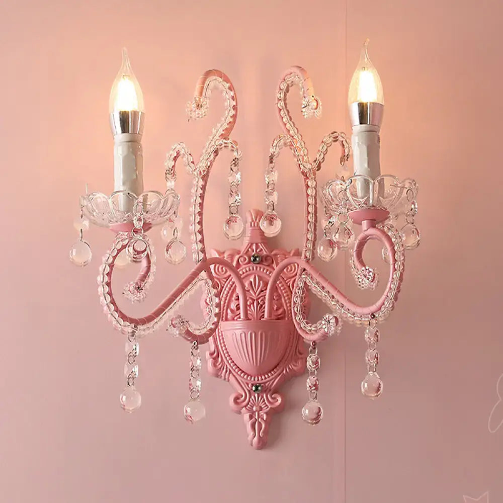 Modern pink metallic scroll arm wall lamp with crystal droplets and two lit candle-style bulbs for bedroom use