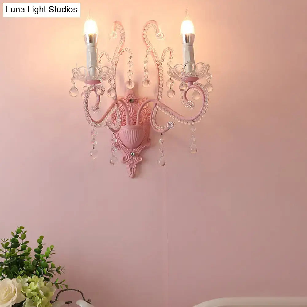 Modern pink metallic scroll arm wall lamp with crystal droplets and two candle-style bulbs for bedroom sconce lighting