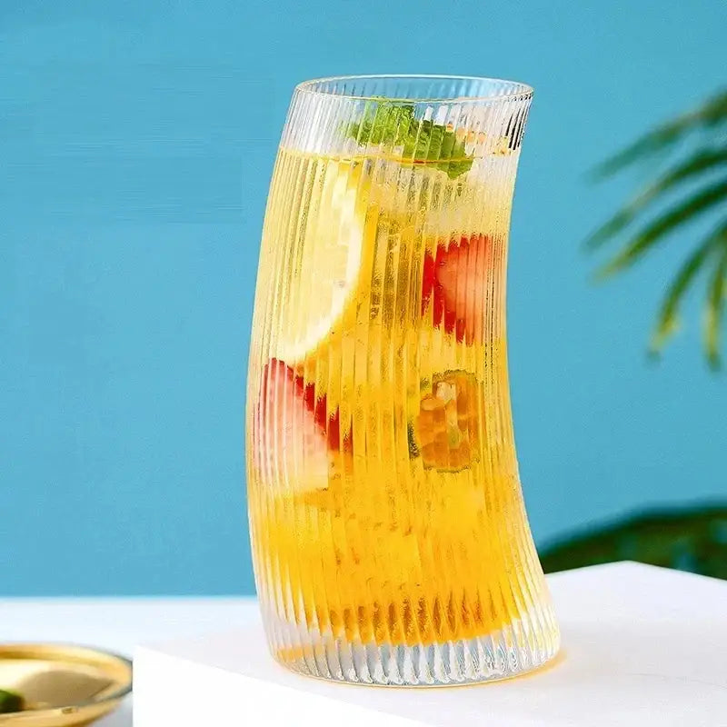A tall modern ribbed sail-shaped glass cup filled with golden beverage and garnished with strawberries lemon and mint leaves