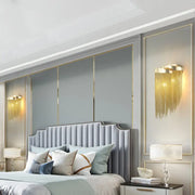 Modern silver and gold tassel design wall lamp with bulb base above a light gray tufted bed with curved headboard and pillows