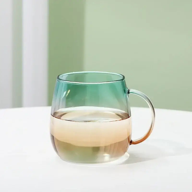A translucent gradient glass mug with teal to amber colors and clear handle from a modern glass teapot set filled halfway with water