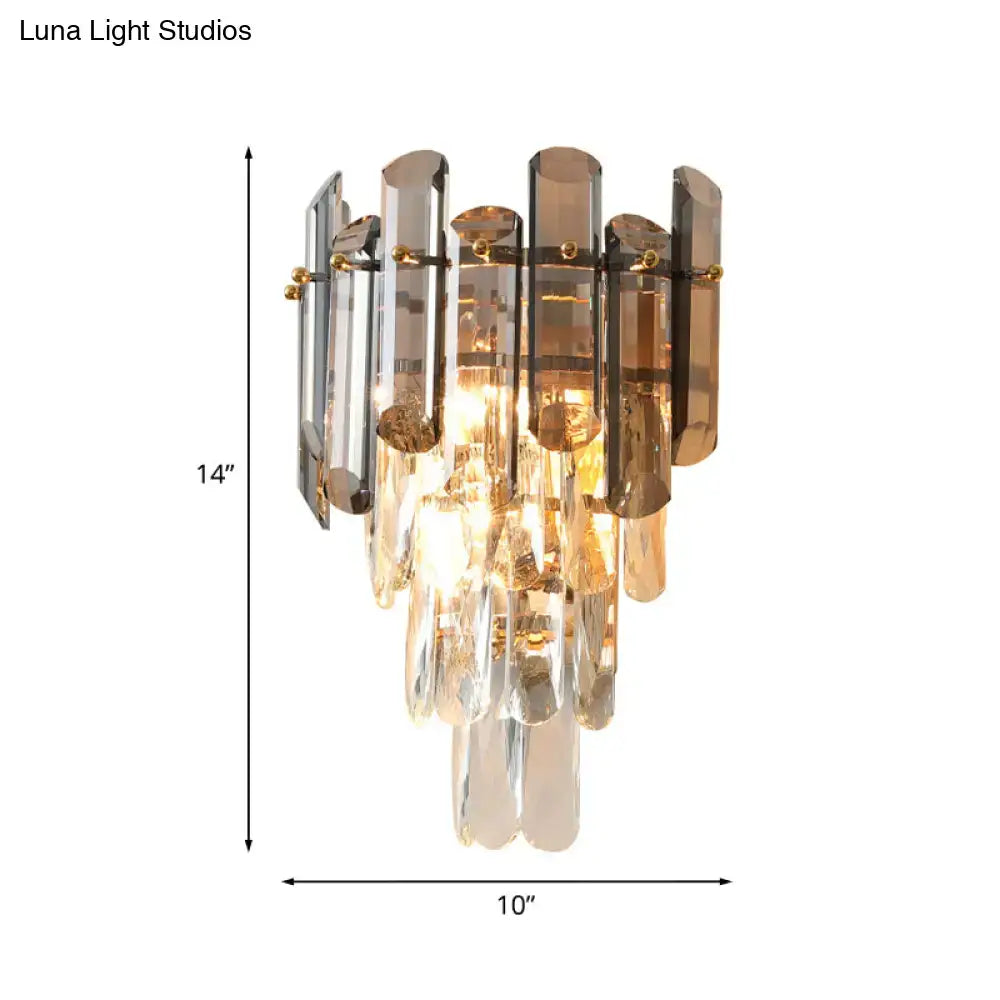A modern tiered clear and smoke grey crystal wall sconce with cascading glass tubes and gold-toned hardware for bedroom lighting