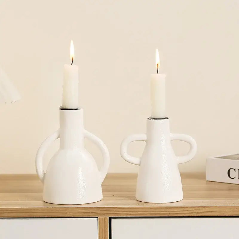 Two white geometric ceramic candle holders with handle grips holding lit white taper candles for decorbites decor