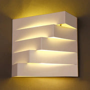 Modern white geometric wall mounted lighting sconce with layered stepped design emitting warm ambient light