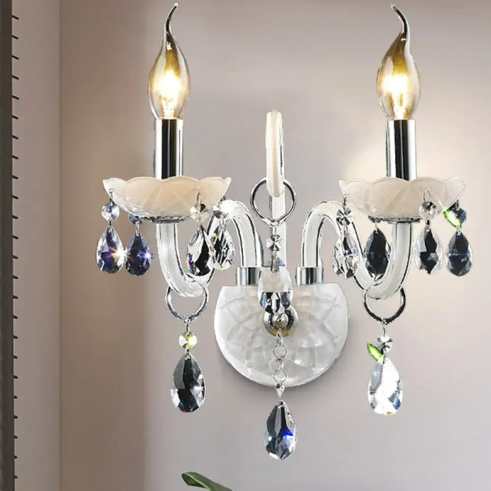 Elegant modern white wall sconce light with teardrop crystal decoration and two lit candle-style bulbs on chrome arms