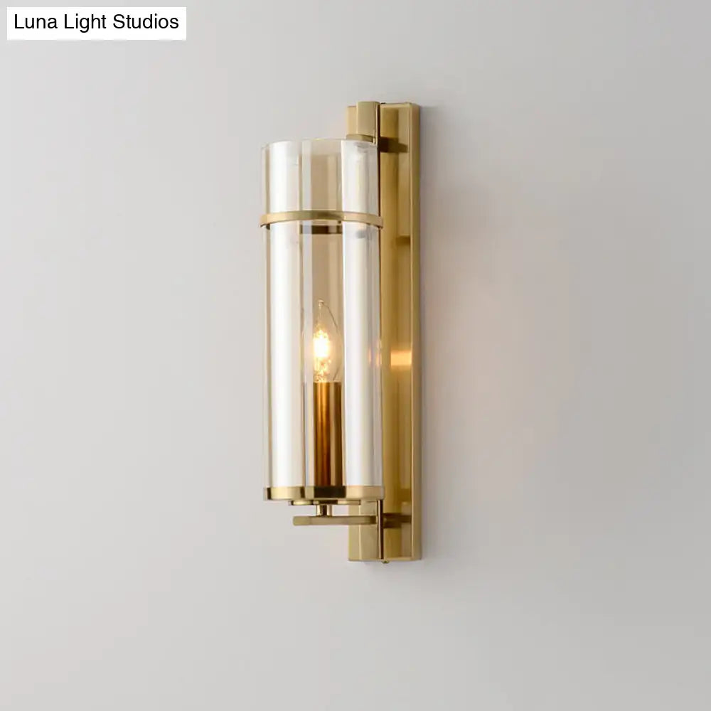 A sleek brass wall lamp with a cylindrical clear glass enclosure and exposed filament bulb for bedroom sconce light