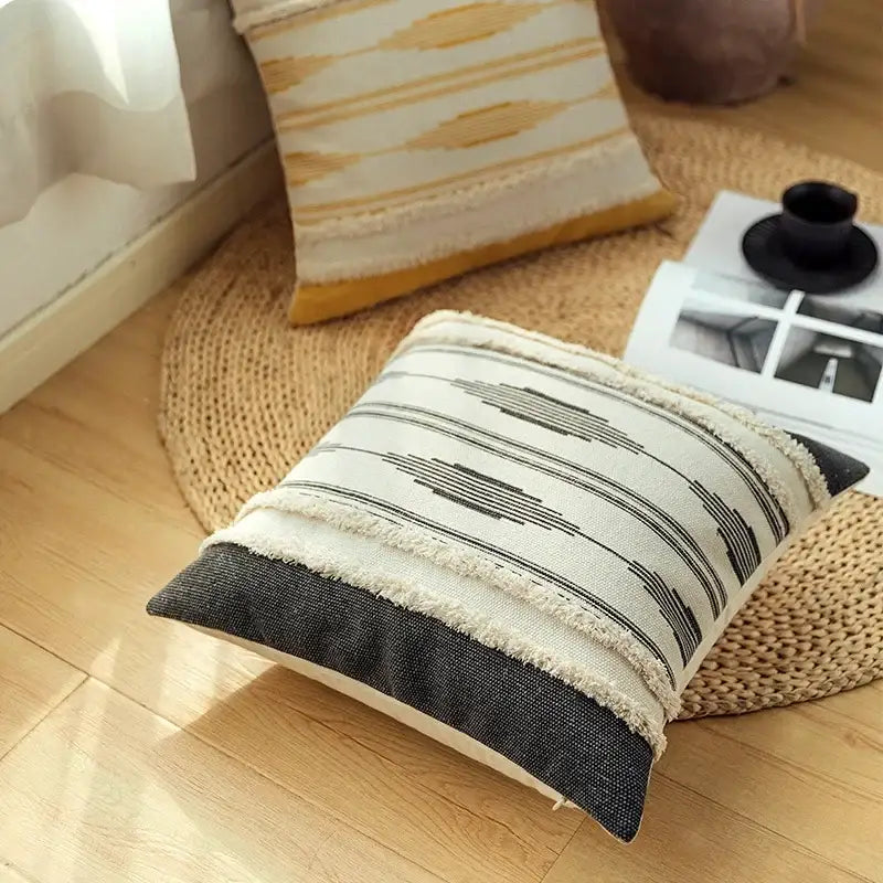 Cream-colored cushion cover set with black geometric stripes and frayed edges on a dark gray base for living room