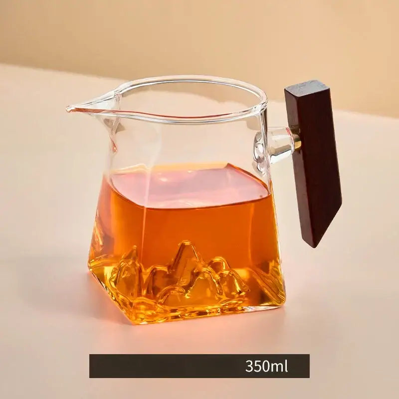 A clear glass tea cup set with a mountain-shaped base and dark wooden handle filled with amber tea for dining room use