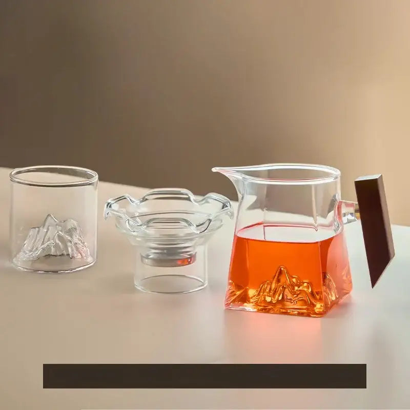 Clear glass teapot with dark wooden handle and mountain-shaped base filled with amber tea in a tea cup set