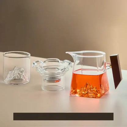 Clear glass teapot with dark wooden handle and mountain-shaped base filled with amber tea in a tea cup set