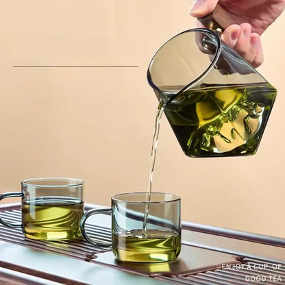 Clear glass tea cup set with hexagonal teapot and gold-toned handle pouring green tea into matching cups
