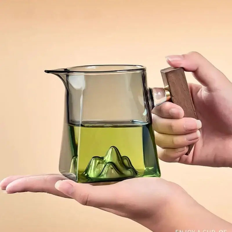 A clear glass tea cup set with wooden handle featuring a green mountain design filled with light green tea